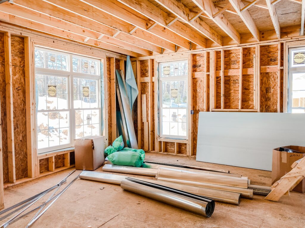 A room under construction with exposed wooden framing, insulation materials, drywall sheets, and building supplies on the floor. Two windows let in natural light—typical of homes rising in new communities St. Cloud is known for.