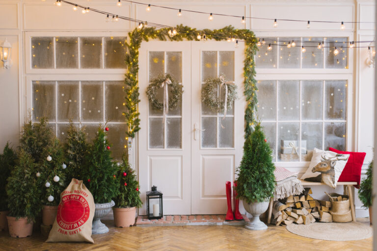 Festive porch with string lights, garland, wreaths on glass doors, potted evergreens, stacked firewood, a holiday sack, and a bench with pillows and a blanket—perfect for celebrating the holidays or to Sell St. Cloud home this festive season.