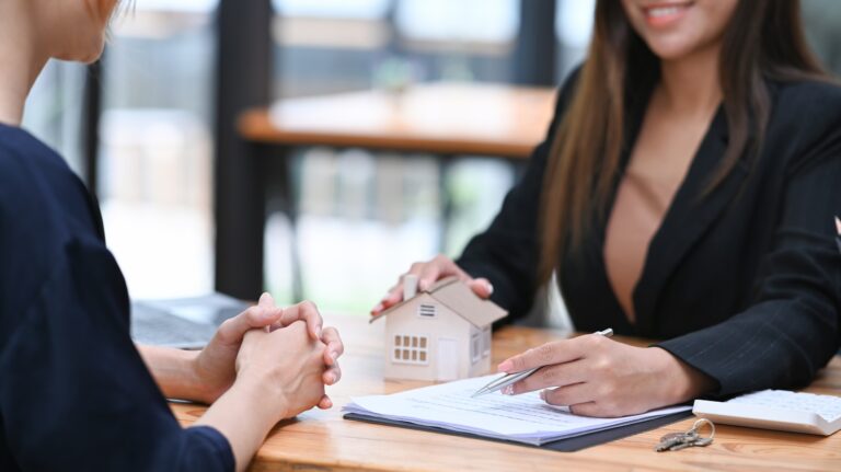 Two women sit at a table in St. Cloud reviewing documents for listing a home. One holds a pen and a small model house, while the other has hands clasped. Keys and a calculator are on the table, hinting at a fresh start this spring.