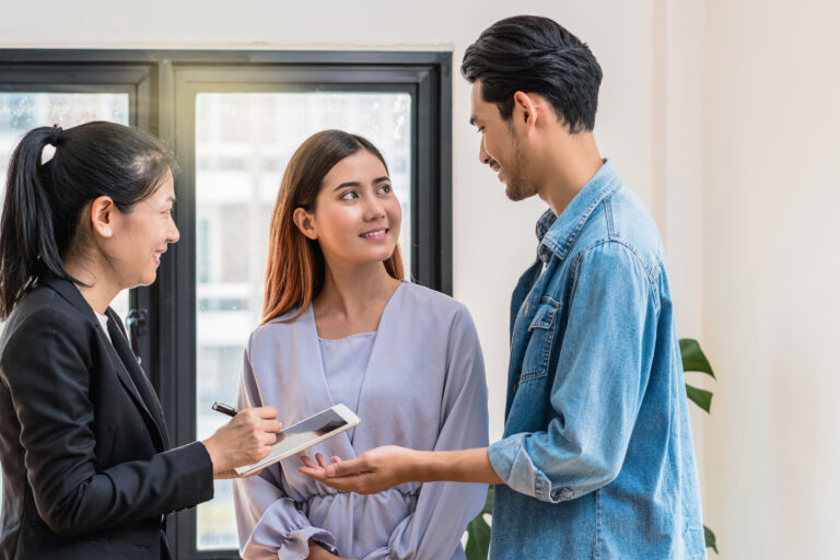 Three people stand near a window; one woman holds a clipboard and pen, consulting with a man and woman who listen attentively, possibly discussing tips for first-time homebuyers buying in St. Cloud, Florida.