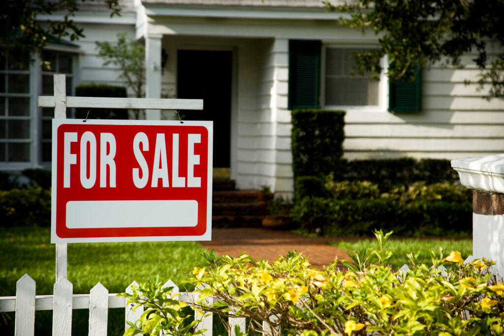 A red and white "For Sale" sign is displayed in front of a white house with green shutters and a lawn, partially visible behind a white picket fence and shrubs—perfect for those searching St. Cloud homes or new real estate opportunities.