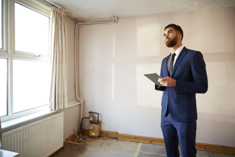 A man in a blue suit holds a tablet and looks up while standing in an empty, unfurnished room—perhaps preparing for a home appraisal or getting ready for selling St. Cloud homes. Bare walls and exposed wires set the scene.