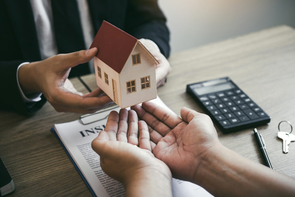 In St. Cloud, a person hands a small model house to another over an insurance document, with keys and a calculator on the table—highlighting how title issues can cause closing delays in real estate transactions.
