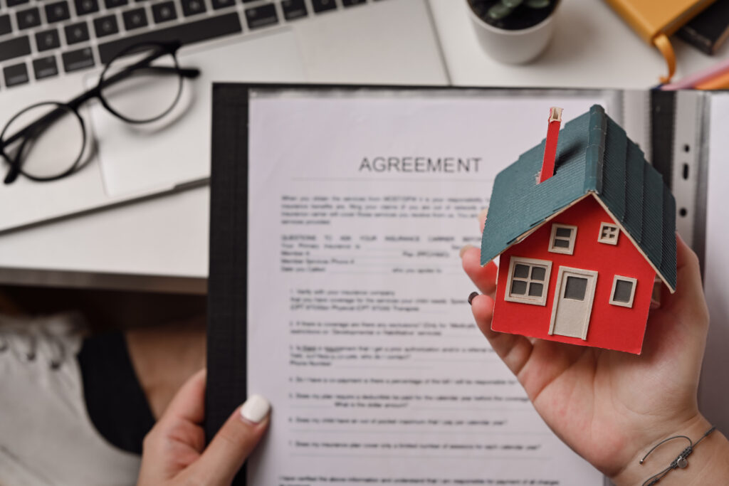 A person holds a small model house and a folder with an agreement document, considering the pros and cons of HOAs, with a laptop, glasses, and a plant visible on the desk.