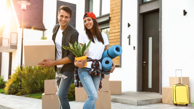 A man and woman, new residents of St. Cloud, stand outside a modern house with moving boxes, a potted plant, yoga mats, and a yellow suitcase, settling in after relocating.