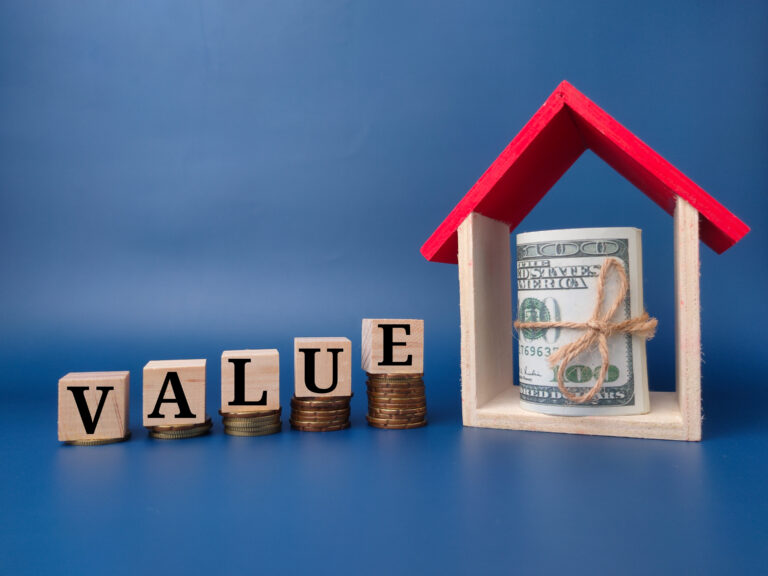 Wooden blocks spelling "VALUE" rest on stacked coins beside a model house containing rolled dollar bills, all set against a blue background, symbolizing the causes of a low appraisal or issues that may impact your home appraisal.