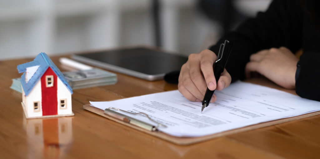 A person signs documents on a clipboard at a desk with a small model house, a tablet, and a stack of cash nearby—an essential step for St. Cloud homebuyers following their home buying guide.