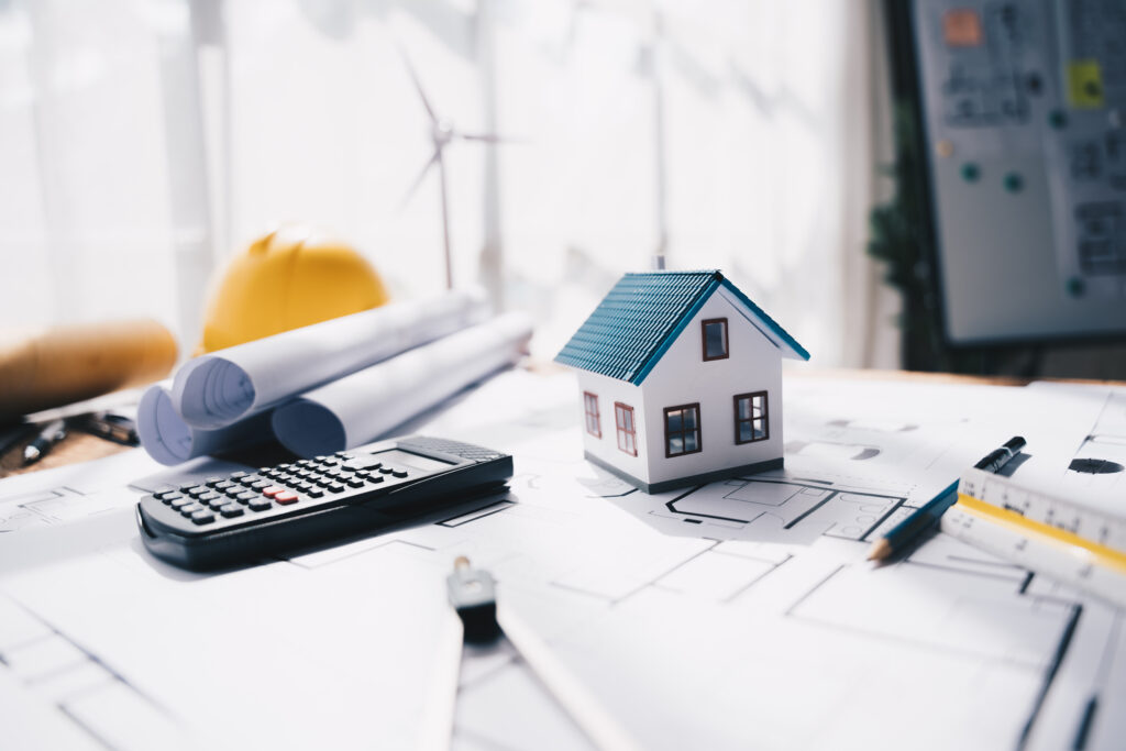 A miniature house, calculator, rolled blueprints, and a yellow hard hat are placed on architectural drawings, illustrating how buyers and sellers in Osceola County can plan for closing costs during their real estate transactions.