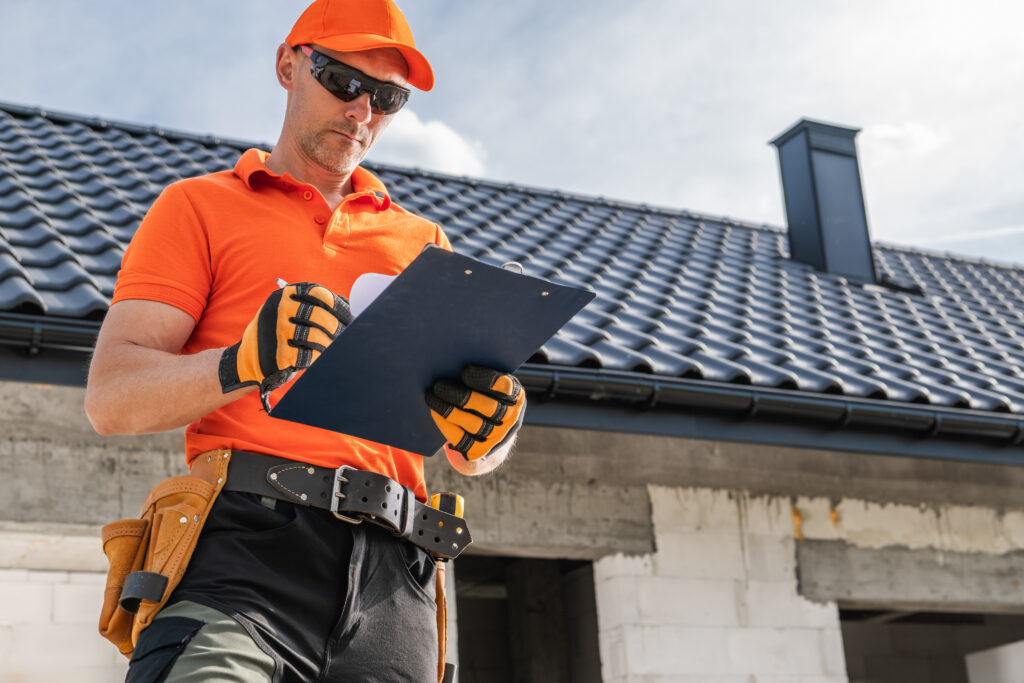 Construction worker in orange shirt and cap writes on a clipboard, preparing for a rainy season home inspection in front of a building with a tiled roof. Tools are visible on his belt.
