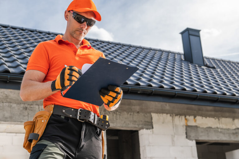 Construction worker in orange shirt and cap writes on a clipboard, preparing for a rainy season home inspection in front of a building with a tiled roof. Tools are visible on his belt.