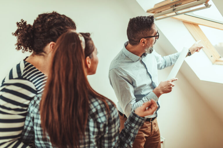 A man holding papers points at a skylight while two buyers stand beside him, looking in the same direction inside a bright room.