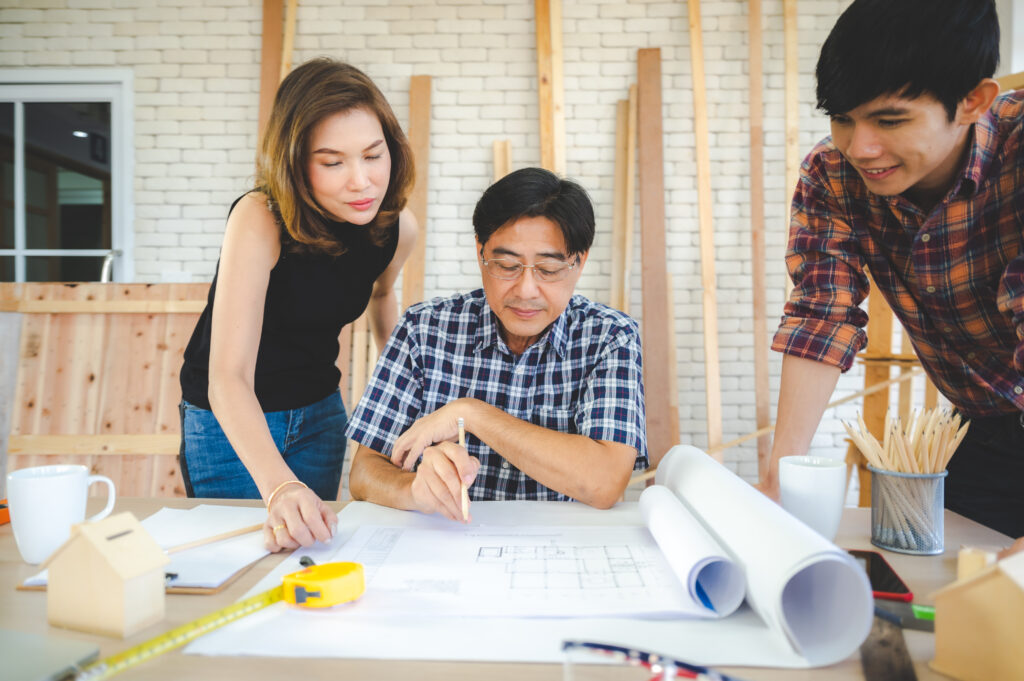 Three people review architectural blueprints on a table, discussing plans to renovate before selling a St. Cloud home, in a workspace filled with wooden planks and tools.