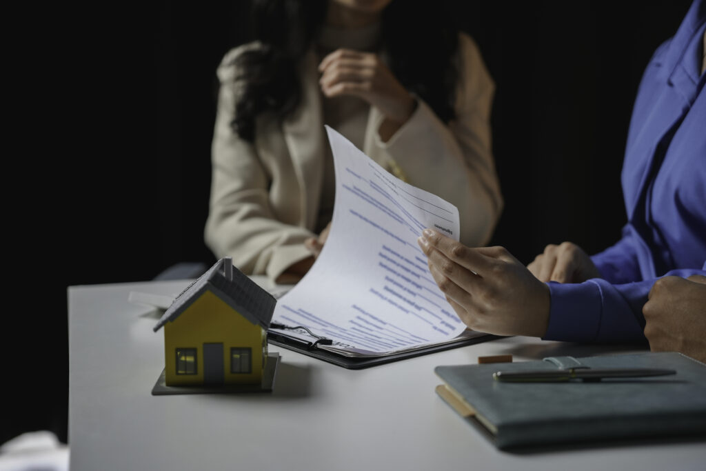 Two people sit at a table with documents, a model house, and writing materials, discussing paperwork related to real estate or property transactions—perhaps reviewing essential details like title insurance, which many St. Cloud homeowners consider when buying or selling their homes.