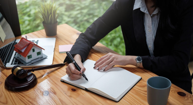 A person in business attire writes in a notebook at a desk with a laptop, house model, gavel, plant, and coffee mug—perhaps preparing documents related to wills and trusts or organizing important probate paperwork.